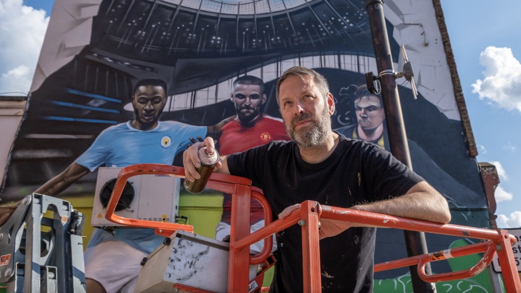 Street artist Jody Thomas poses paint can in hand in front of his football mural