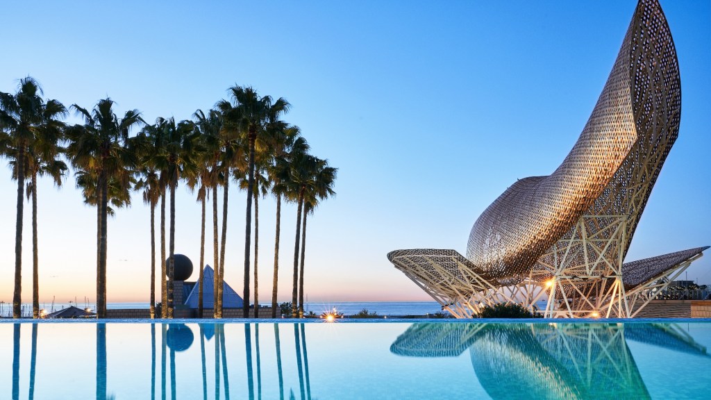 Frank Gehry’s El Peix goldfish structure looks out to sea from an infinity pool. The skies are blue, and palm trees reflect in the pool