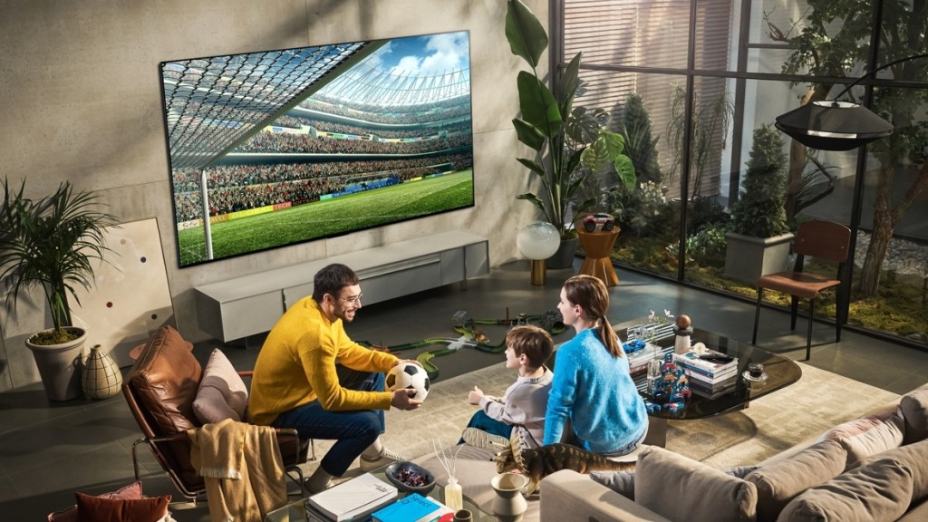 A family appear to be watching a football match on the giant 97inch LG G2 OLED, while holding a football, as you do