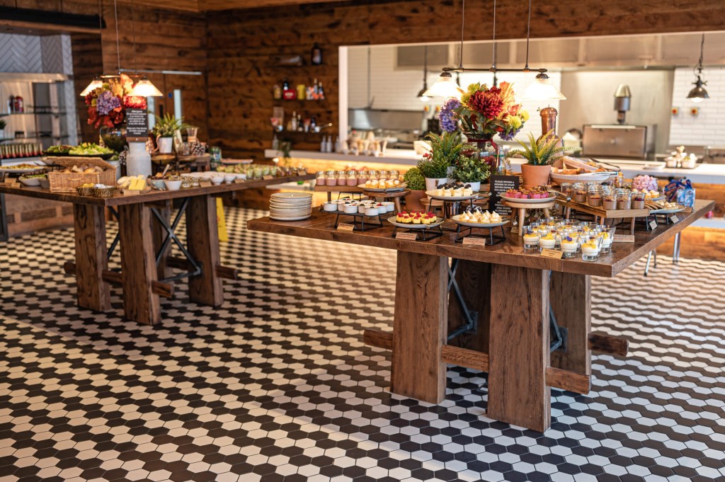 A decorated buffet table filled with an array of desserts and appetizers, featuring colorful pastries, cakes, and small dishes, set against a wooden backdrop and patterned floor.
