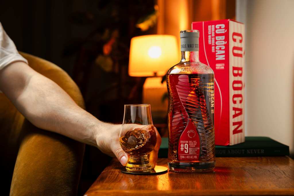 A hand reaching for a glass of whiskey on a wooden table, with a decorative bottle of Cù Bòcan whiskey and a softly lit lamp in the background.