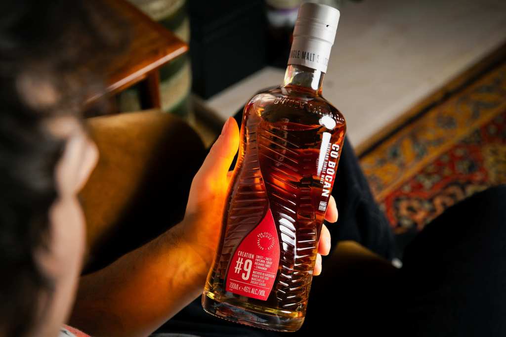 A person holding a bottle of whiskey with a red label, sitting in a cozy setting with a patterned rug in the background.