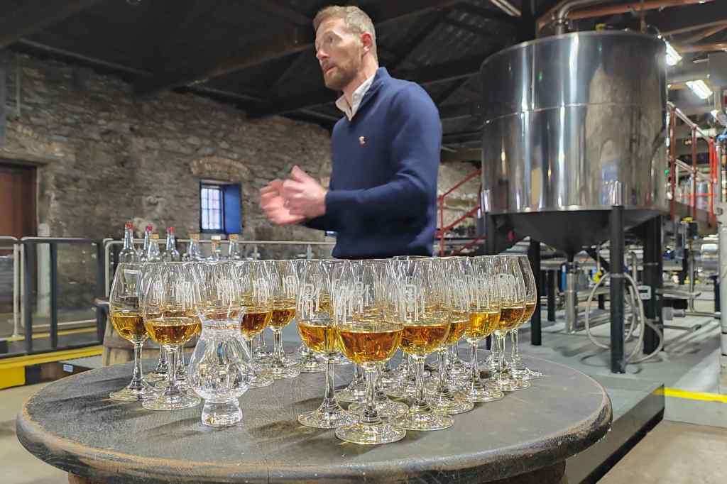 A distillery setting featuring a man gesturing while speaking, with a row of filled glasses of whiskey on a wooden barrel in the foreground.