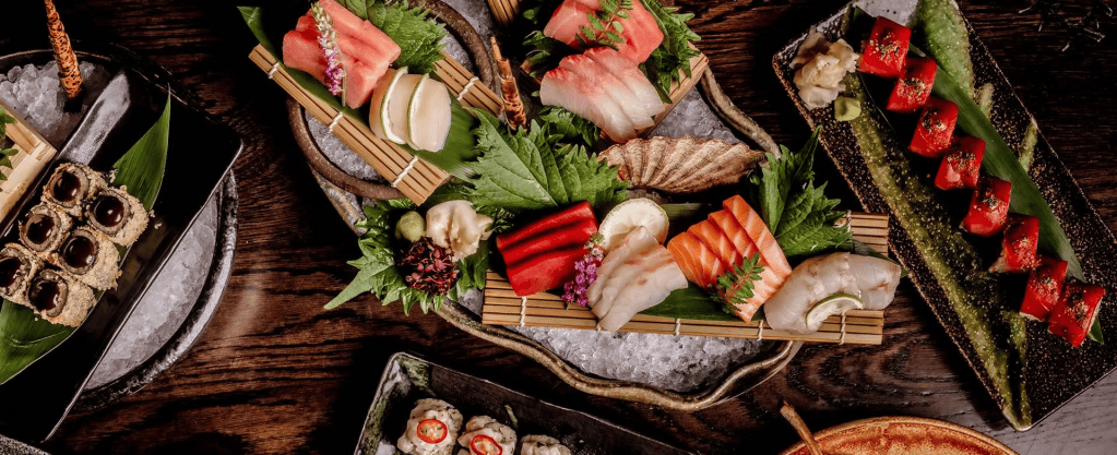 A beautifully arranged sushi platter featuring various types of sushi, sashimi, and garnishes on wooden and stone plates, presented on a bed of ice.