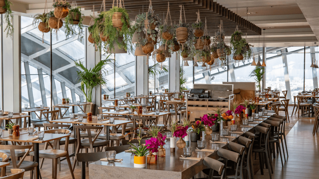 A modern restaurant interior featuring wooden tables and chairs, adorned with colorful floral arrangements and hanging plants, with large windows providing a view of the exterior.