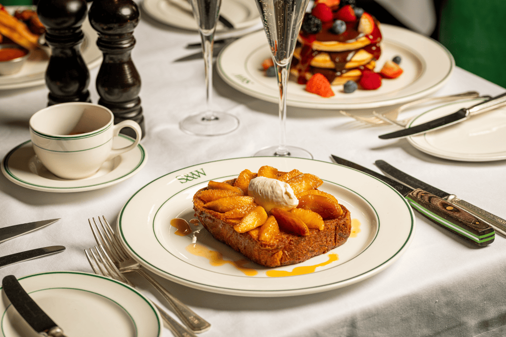 A close-up of a plate featuring a slice of warm bread topped with caramelized apples and a dollop of whipped cream, accompanied by a cup and saucer, and a glass of sparkling wine on a dining table set with utensils.