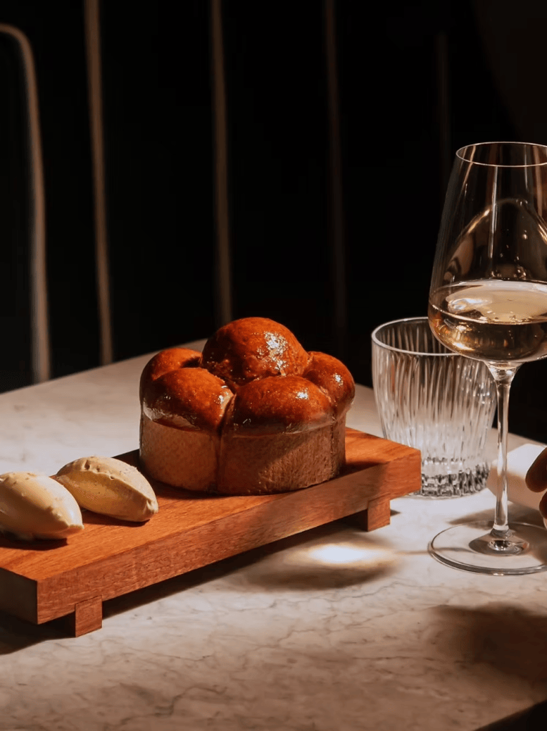 A freshly baked round bread served on a wooden board alongside two pieces of butter and a glass of white wine on a marble table.