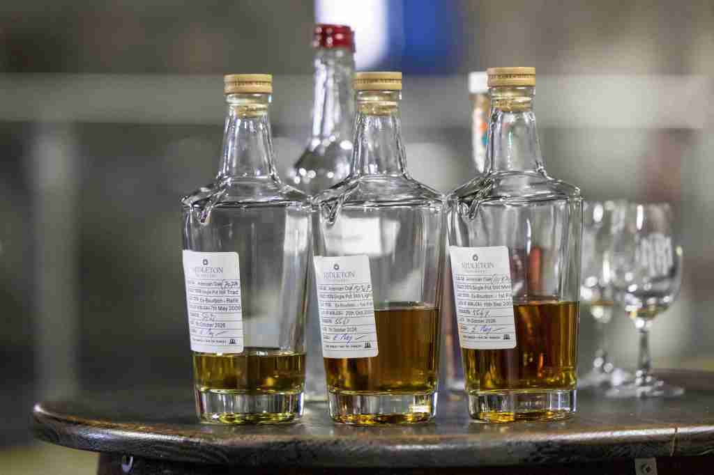 Three glass bottles of whiskey with labels, displayed on a wooden surface, with a few empty glasses in the background.
