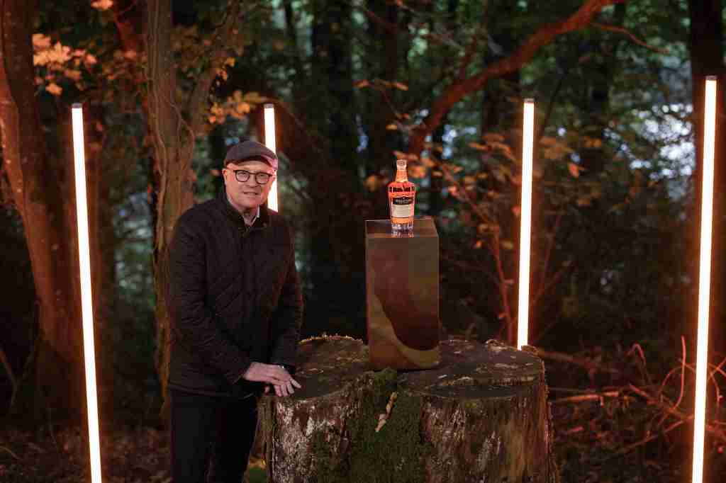 A person wearing glasses and a cap stands next to a tree stump with a bottle of whiskey on a pedestal, surrounded by illuminated vertical lights in a forest setting.