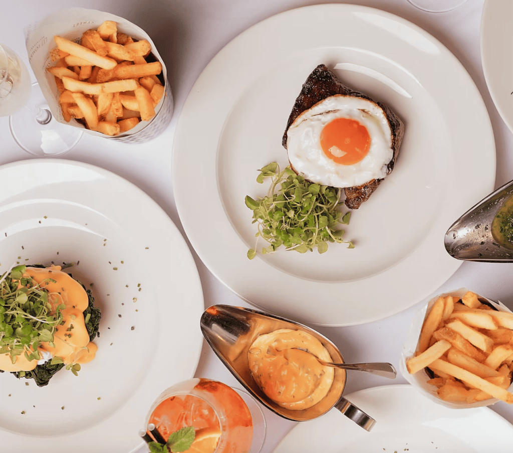 A top-down view of a dining table with various dishes including a piece of steak topped with a fried egg, a serving of fries, a creamy sauce in a bowl, and a side of spinach with hollandaise sauce. A cocktail is also visible in the foreground.