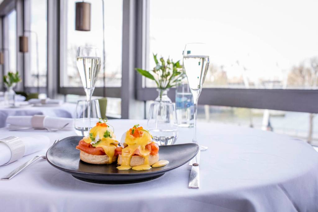 A gourmet plate of eggs Benedict on a black dish, topped with salmon and hollandaise sauce, served on a white tablecloth alongside a flute of champagne and a glass of water, with a scenic background visible through large windows.